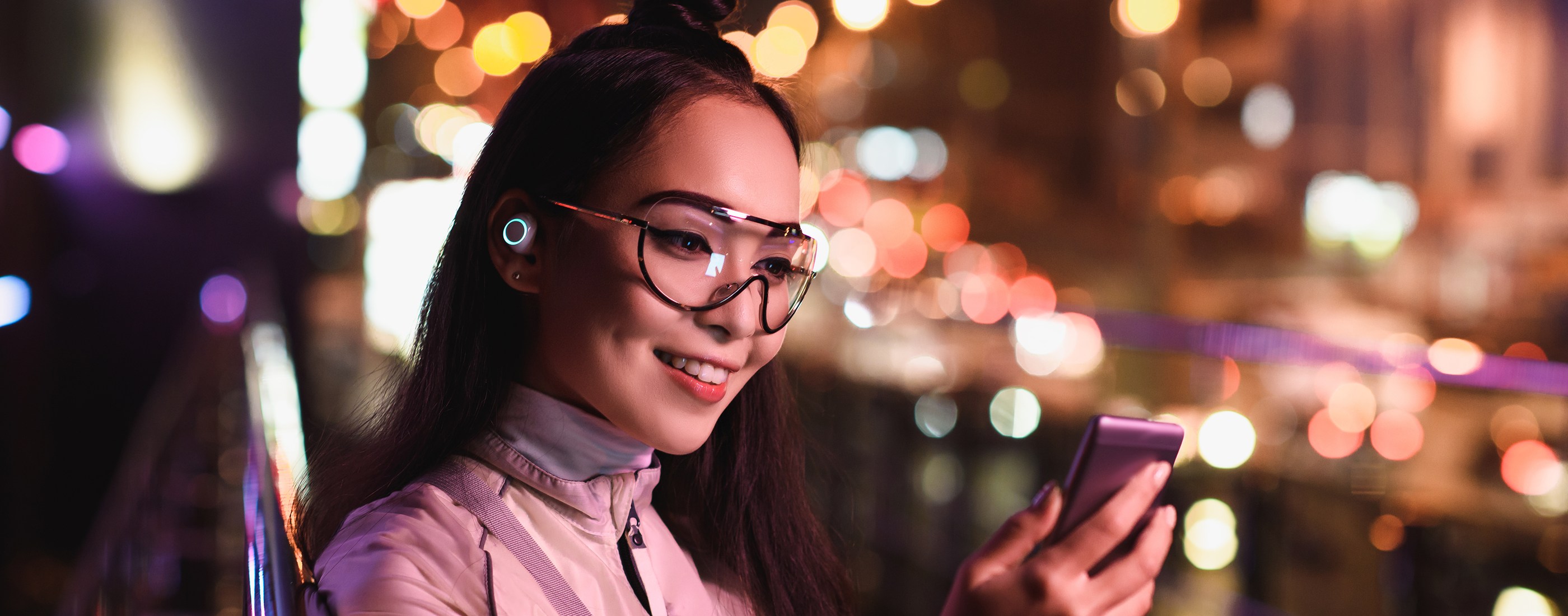 A woman with an earbud smiling at her phone on a city street at night, glowing city lights behind her
