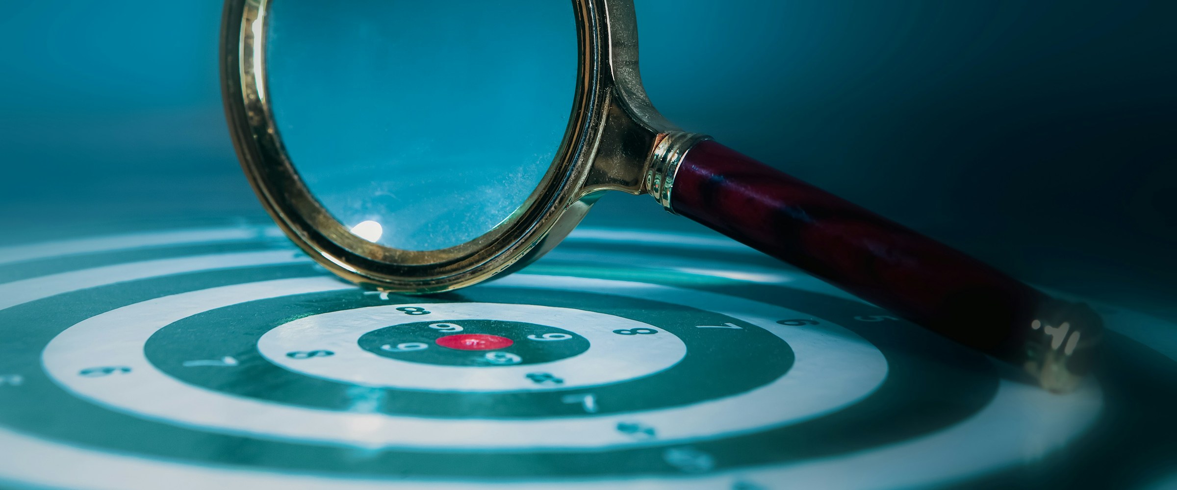A magnifying glass resting over the bullseye of a target, lit in deep teal