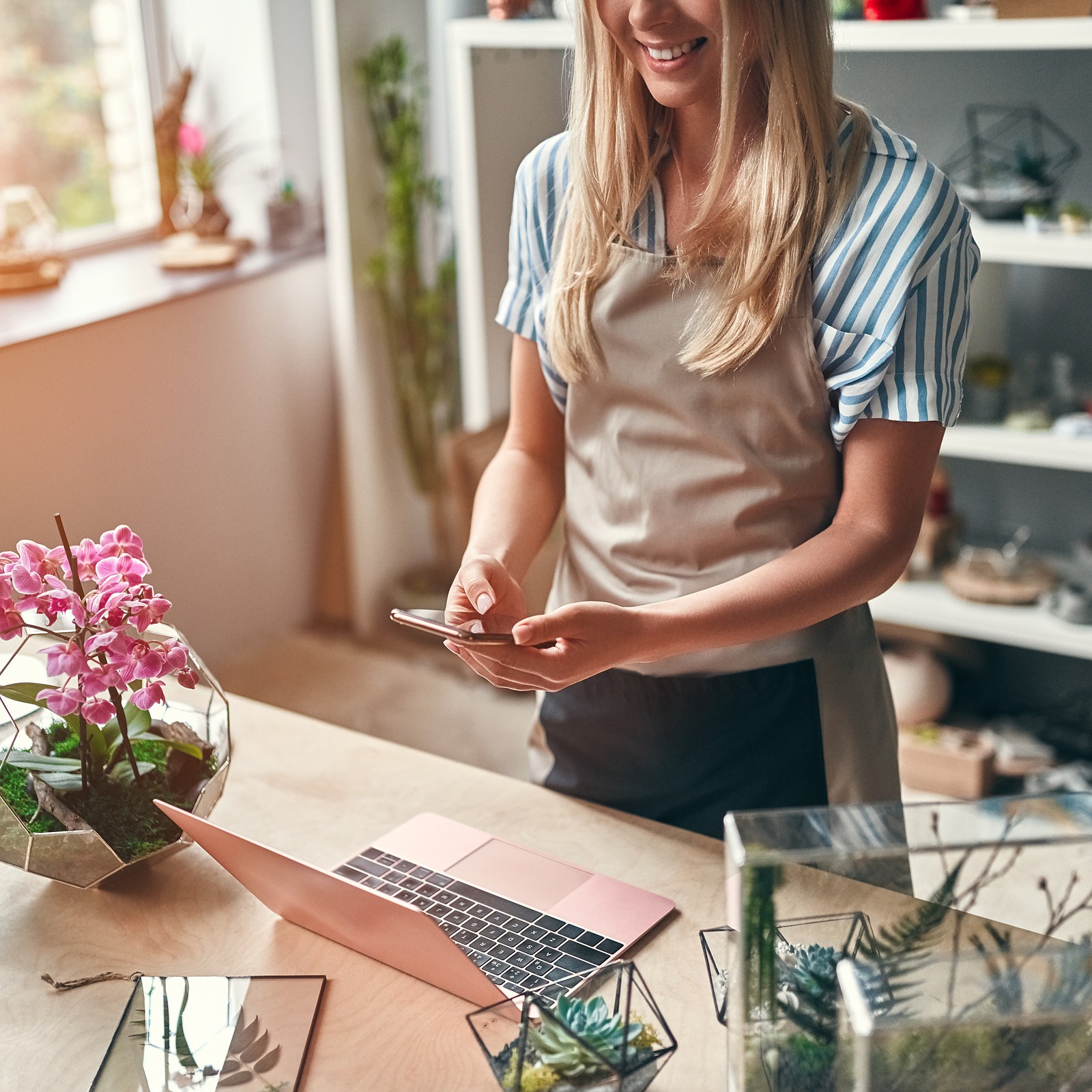 A flower shop owner checking her phone at the counter beside orchids and her laptop